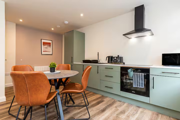 Modern, conveneient kitchen and dining area including four chairs, round table with artificial plant, extractor fan, oven, microwave, toaster, and kettle